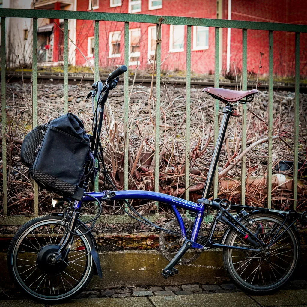 Brom-E quer am Brompton Electric, abgestellt vor einem Zaun
